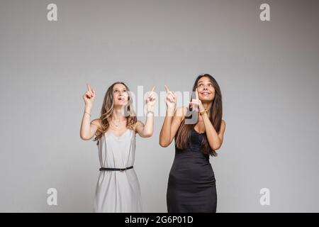 Zwei hübsche Frauen, die im Studio stehen und ihre Finger nach oben zeigen, isoliert auf grauem Hintergrund. Party-Konzept. Speicherplatz kopieren Stockfoto