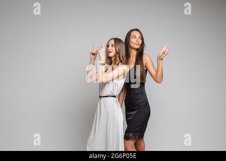 Zwei hübsche Frauen, die im Studio stehen und ihre Finger nach oben zeigen, isoliert auf grauem Hintergrund. Party-Konzept. Speicherplatz kopieren Stockfoto