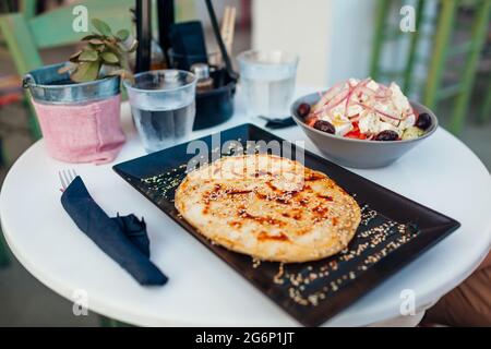 Griechische weiche Pita, gefüllt mit Käse, bestreut mit Sesamsamen, Honig, serviert mit griechischem Salat und Wasser im Café Stockfoto