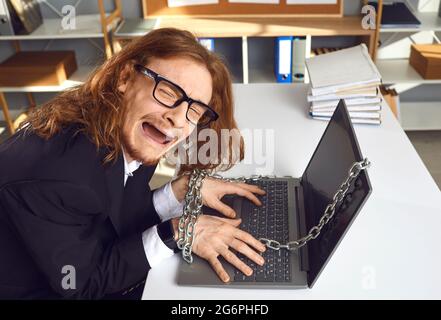 Müder Büroangestellter, der am Schreibtisch saß, war an einen Laptop gekettet und weinte, weil er nicht nach Hause gehen konnte Stockfoto