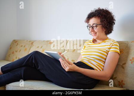 Die junge Frau greift auf ihr digitales Tablet, das auf dem Sofa sitzt Stockfoto