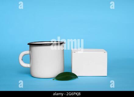 Ein Becher aus weißem Metall, eine weiße Schachtel und ein grünes Ficusblatt auf blauem Hintergrund. Stockfoto