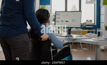 Ein Geschäftsmann hilft seinem behinderten schwarzen Kollegen, der in das Firmenbüro eintritt und den Rollstuhl in die Nähe des Schreibtisches schiebt. Afrikanisch behinderte gelähmte Frau begrüßt Kollegen, die bei einem Finanzprojekt arbeiten Stockfoto