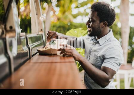 Junger afrikanischer Mann, der im Food-Truck-Schalter im Freien im Stadtpark isst - Fokus auf Gesicht Stockfoto