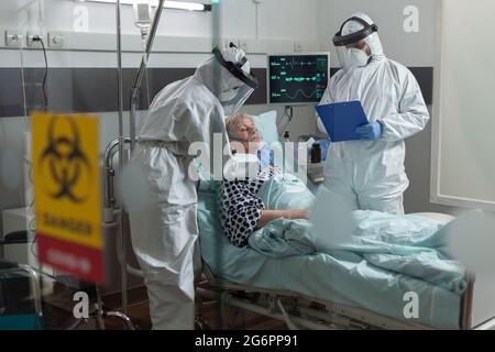 Medizinische Krankenschwester in PSA-Anzug, die im Zuge einer globalen Pandemie mit Coronavirus eine Sauerstoffmaske an einen älteren Patienten legt, und der Arzt notiert sich in der Zwischenablage. Stockfoto