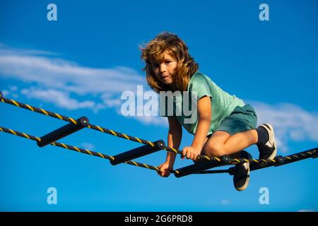 Kind klettert im Netz. Netter Junge klettert die Leiter auf dem Spielplatz hoch. Kind klettert die Leiter gegen den blauen Himmel hinauf. Stockfoto