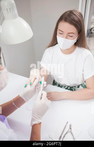 Vertikale Aufnahme einer jungen Frau mit Gesichtsmaske, die während einer Coronavirus-Pandemie im Salon Maniküre erhält Stockfoto