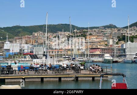 Menschen, die in einem Restaurant im Freien am Meer am Alten Hafen (Porto Antico) von Genua mit der Stadt im Hintergrund im Sommer zu Mittag essen, Italien Stockfoto