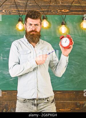 Der Lehrer in der Brille hält Wecker und Stift. Bärtiger Hipster hält die Uhr, Kreidetafel auf dem Hintergrund. Mann mit Bart und Schnurrbart im strengen Gesicht Stockfoto