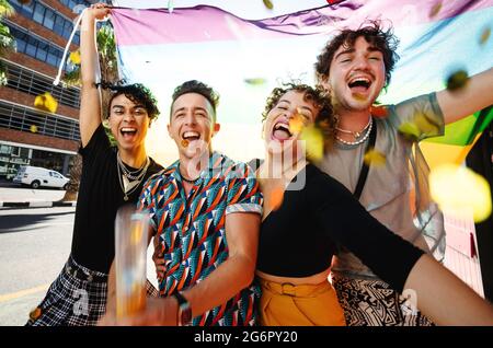 Gruppe von nicht-konformen jungen Menschen, die mit der Flagge des Regenbogens feiern. Vier queere Menschen lächeln fröhlich während der Feier Stockfoto