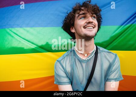 Sorgloser schwuler Mann, der sich seiner Identität annimmt. Fröhliche junge Homosexuell lächeln fröhlich, während sie gegen eine stolze Flagge stehen. Junger queerer Mann mit Lidschatten aga Stockfoto