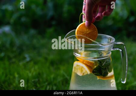 Defocus Hand hält Dummy Zitrone hängen über Glas Krug Limonade mit Scheibe Zitrone und Blätter der Minze auf natürlichem grünem Hintergrund. Krug des Sommers Stockfoto