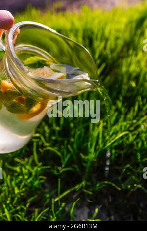 Unschärfe-Hand hält Glaskrug Limonade mit Zitrone und Minze auf natürlichem grünen Hintergrund. Krug mit einem kühlen Sommercocktail oder Punsch im Freien, outsi Stockfoto