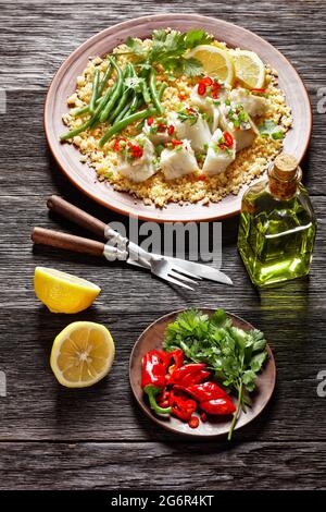 Bacalhau com todos, Kabeljau mit Bulgur, gedämpfte grüne Bohnen, bestreut mit scharfer Chilischote und Frühlingszwiebeln auf einem rustikalen Teller, auf einem Holztisch, por Stockfoto