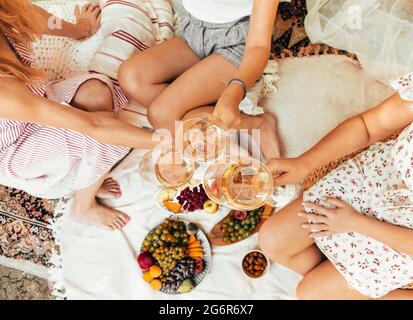 Von oben anonymen Freundinnen klirren Gläser Wein, während sie auf Decken neben Tellern mit Früchten während des Picknicks sitzen Stockfoto