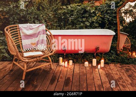 Brennende Kerzen und Korbsessel mit Buch und Wein auf Holzweg in der Nähe von Badewanne und Spiegel im Garten platziert Stockfoto