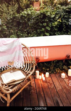 Brennende Kerzen und Korbsessel mit Buch und Wein auf Holzweg in der Nähe von Badewanne und Spiegel im Garten platziert Stockfoto