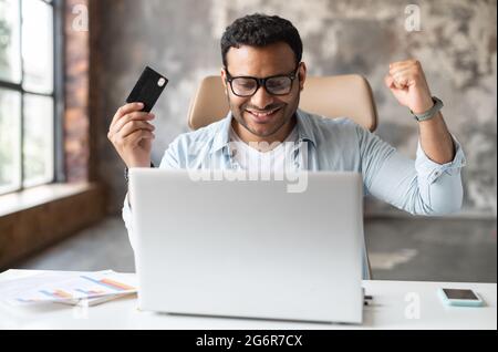 Ein Mann mit gemischter Rasse kaufte online lang erwartete Einkäufe, saß am Schreibtisch und hob die Fäuste in einer triumphalen Geste, hielt die Kreditkarte, ein indischer Mann blickt auf den Laptop-Bildschirm und freut sich Stockfoto