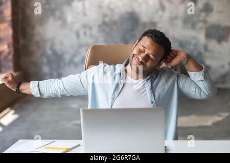 Fröhlicher indischer Büroangestellter, der die Arme streckt am Schreibtisch sitzt, ruht sich nach produktiver Arbeit mit dem Laptop oder nach dem fertigen Projekt aus, macht Pause und fühlt sich zufrieden Stockfoto