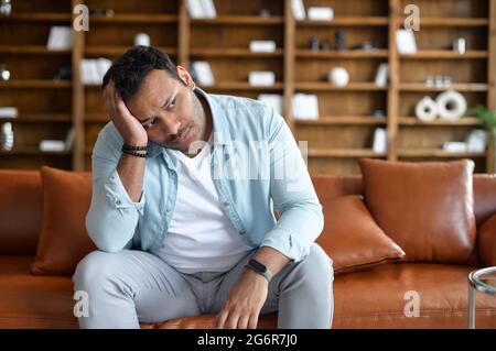 Frustrierter indischer Mann fühlt Zweifel und Verzweiflung, sitzt mit verärgerter Gesichtsausdruck auf dem Sofa und hält den Kopf, östlicher Kerl fühlt Kopfschmerzen und Depressionen, leidet an Reue Stockfoto