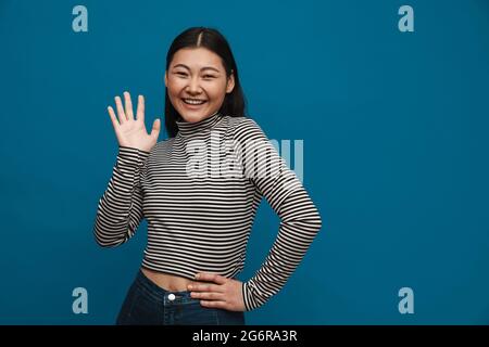 Porträt einer glücklichen beiläufigen jungen asiatischen Frau, die über blauem Wandhintergrund mit winkender Hand steht Stockfoto