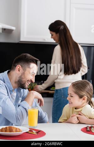 Glücklicher Vater und Tochter lachen beim Frühstück in der Küche in der Nähe verschwommene Mutter Stockfoto