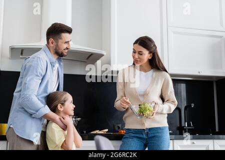 Lächelnde Frau hält eine Schüssel mit Gemüsesalat in der Nähe von Ehemann und Ehefrau in der Küche Stockfoto