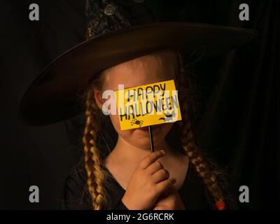 Kleines hübsches Mädchen Hexe in schwarzem Zauberer Hut hält Gruß Happy Halloween und lacht. Lächelndes Kind in Geisterkostüm. Festliche Herbstferien Stockfoto