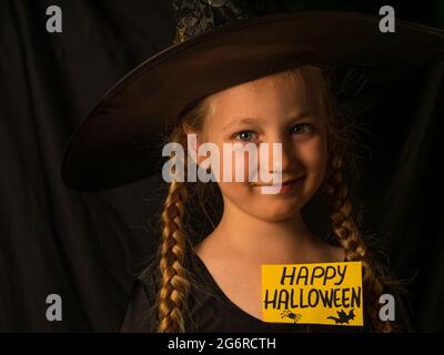 Kleines hübsches Mädchen Hexe in schwarzem Zauberer Hut hält Gruß Happy Halloween und lacht. Lächelndes, positives Kind in Geisterparty-Kostüm gekleidet Stockfoto