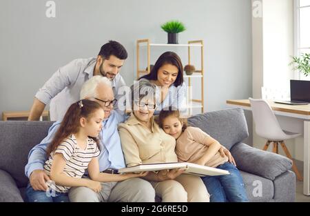 Fröhliche Familie, die auf einem Sofa sitzt, ein Buch liest oder gemeinsam durch ein Fotoalbum schaut Stockfoto