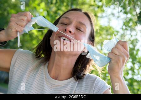 Freudige Frau reißt die medizinische Maske auf ihr Gesicht, sie lächelt und fühlt sich frei. Konzept der Bekämpfung des Virus, das Ende der Pandemie. Stockfoto