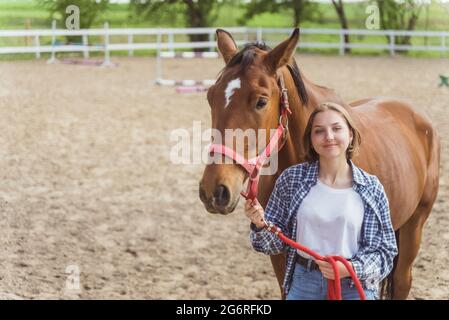 Junges Mädchen mit ihrem Bay-Pferd im Stall. Mädchen hält Pferd Bleiseil Blick auf sie mit Liebe. Posiert für die Kamera. Sandarena mit Holzzaun im Hintergrund. Stockfoto