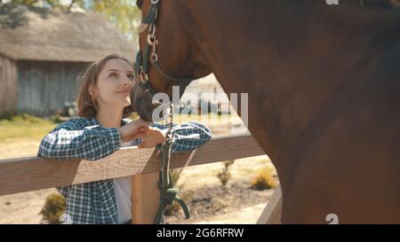 Junges Mädchen, das mit Liebe auf ihr Dark Bay Pferd schaut. Mädchen, das ihre Hände auf den Holzzaun gelegt hat. Rückansicht eines braunen Hengstes. Pferdefarm und Holzstall im Hintergrund. Stockfoto