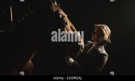 Weibliche Pferdebesitzerin mit ihrem Dark Bay Horse auf schwarzem Hintergrund. Blonde Mädchen trägt Handschuhe und in einem schwarzen Mantel Befestigung Pferd Halfter gekleidet. Reiten zur freien Verfügung. Stockfoto