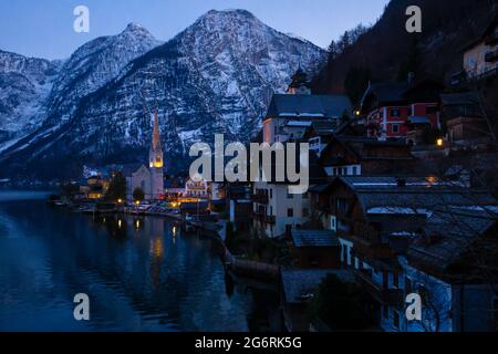 Blick auf den schönen Hallstätter See und die berühmte Kirche in letzter Zeit Abend nach Sonnenuntergang im frühen Frühling mit bedeckten Bergketten In etwas Schnee im Hintergrund Stockfoto