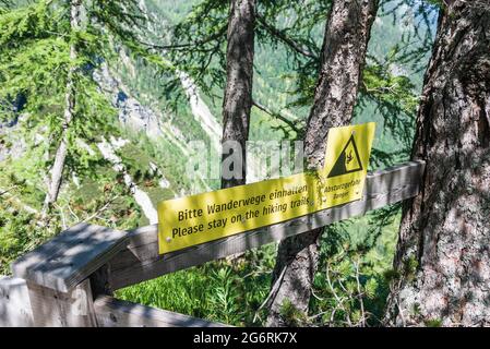 Das gelbe Warnschild, das von Bäumen mit schwarzen Buchstaben auf deutsch und englisch auf dem Wanderweg beleuchtet wird. Bitte Wanderwegen halten, absturzgefa Stockfoto