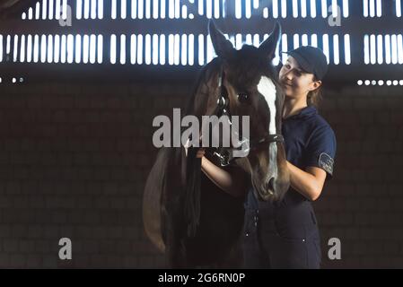 Weibliche Jockey mit ihrem Seal braunen Pferd im Stall. Frau hält das Bleiseil des Pferdes und lächelt es an. Ihre Zuneigung zum Ausdruck bringen. Reiten zur freien Verfügung. Stockfoto