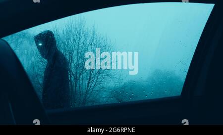 Ein Horrorkonzept. Durch ein Autofenster auf ein unheimliches übernatürliches Wesen mit leuchtenden Augen blicken, das an einem Auto vorbeiläuft. An einem launischen Winterabend. Stockfoto