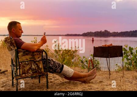 Mann mit Handy in der Hand genießt die herrliche Landschaft, während er am Strand sitzt und einen Grill grilt Stockfoto