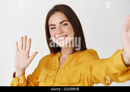 Junge Frau, die Selfie macht und sich selbst auf dem Smartphone aufzeichnet. Frau winkt während eines Videoanrufs mit der Hand auf die Kamera und steht auf weißem Hintergrund Stockfoto