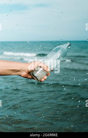 Ein kaukasischer Mann gießt Wasser aus einer wiederverwendbaren Glasflasche vor dem Ozean Stockfoto