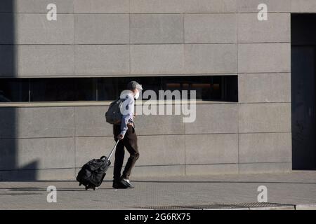 Mann mit Maske, Mütze, Koffer auf Rädern, Rucksack, der die Straße entlang in der Nähe einer grauen Wand geht. Stockfoto