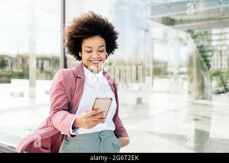Glückliche schwarze Geschäftsfrau, die ihr Mobiltelefon im Freien verwendet. Sie trägt formelle Kleidung und benutzt Kopfhörer. Stockfoto