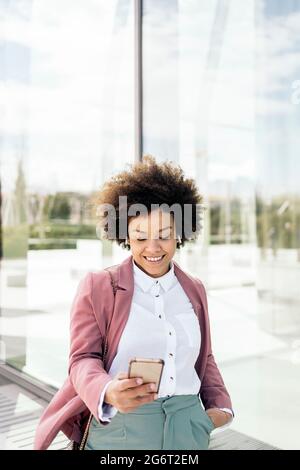 Glückliche schwarze Geschäftsfrau, die ihr Mobiltelefon im Freien verwendet. Sie trägt formelle Kleidung und benutzt Kopfhörer. Stockfoto