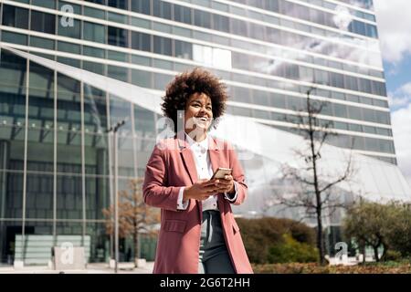 Glückliche schwarze Geschäftsfrau mit afro-Haaren lächelnd und mit ihrem Mobiltelefon im Freien in einem Geschäftsviertel. Stockfoto