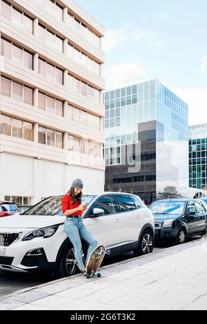 Glückliche junge Skaterin, die in einem Auto auf der Straße sitzt und ihr Handy benutzt. Stockfoto