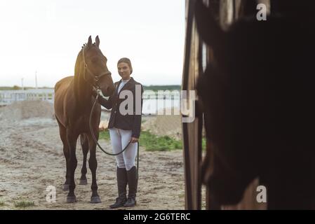 Pferdefrau, die mit ihrem dunklen Bay-Pferd vor dem Stall steht. Posiert für die Kamera. Blick von der Tür des Stalls. Pferdefarm mit Holzzaun im Hintergrund. Tagesaufnahme. Stockfoto