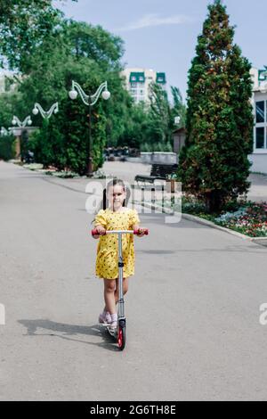 Ein süßes Mädchen in einer gelben Sundress lernt, mit einem Roller durch die Stadt zu fahren und genießt einen heißen Sommertag Stockfoto