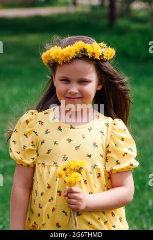 Der Wind bläst die Haare eines Mädchens in einem gelben Kleid und einen Kranz aus gelben Dandelionen auf ihren Kopf im Frühlingsshof Stockfoto