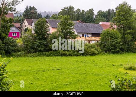 Zwei Arbeiter bauen das Dachgebinde einer Scheune in Görsdorf, einem Weiler der Stadt Schalkau in Thüringen, Deutschland. Im Hintergrund stehen halbverputzte und verputzte Häuser des Dorfes. Während der deutschen Teilung lag Görsdorf innerhalb der 500-Meter-Schutzzone der DDR und wurde 1981 von einer etwa 3,5-Meter-Betonmauer umgeben, die als Sichtschutz diente. Stockfoto